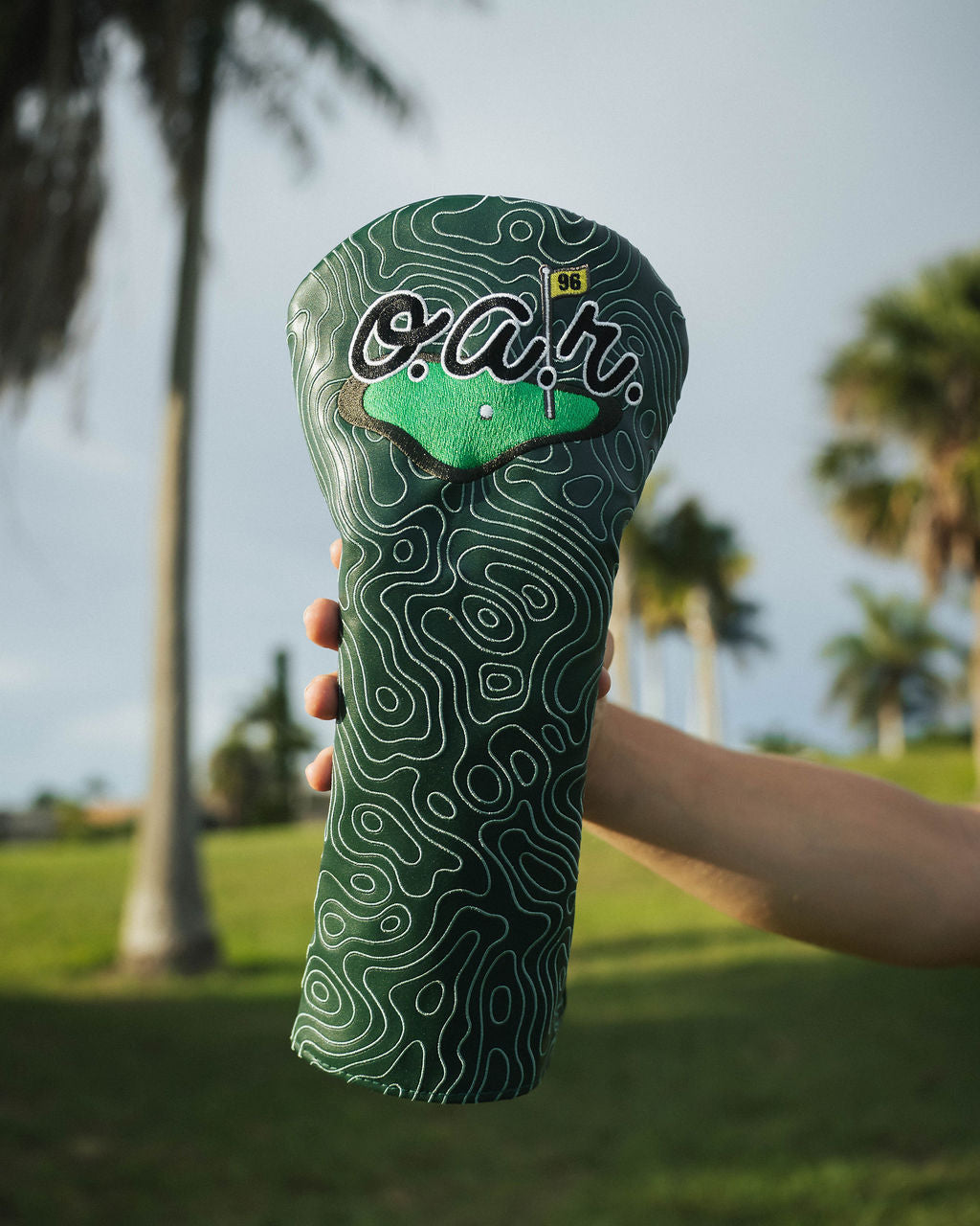 Green golf club head cover with 'o.a.r.' branding held in front of palm trees.