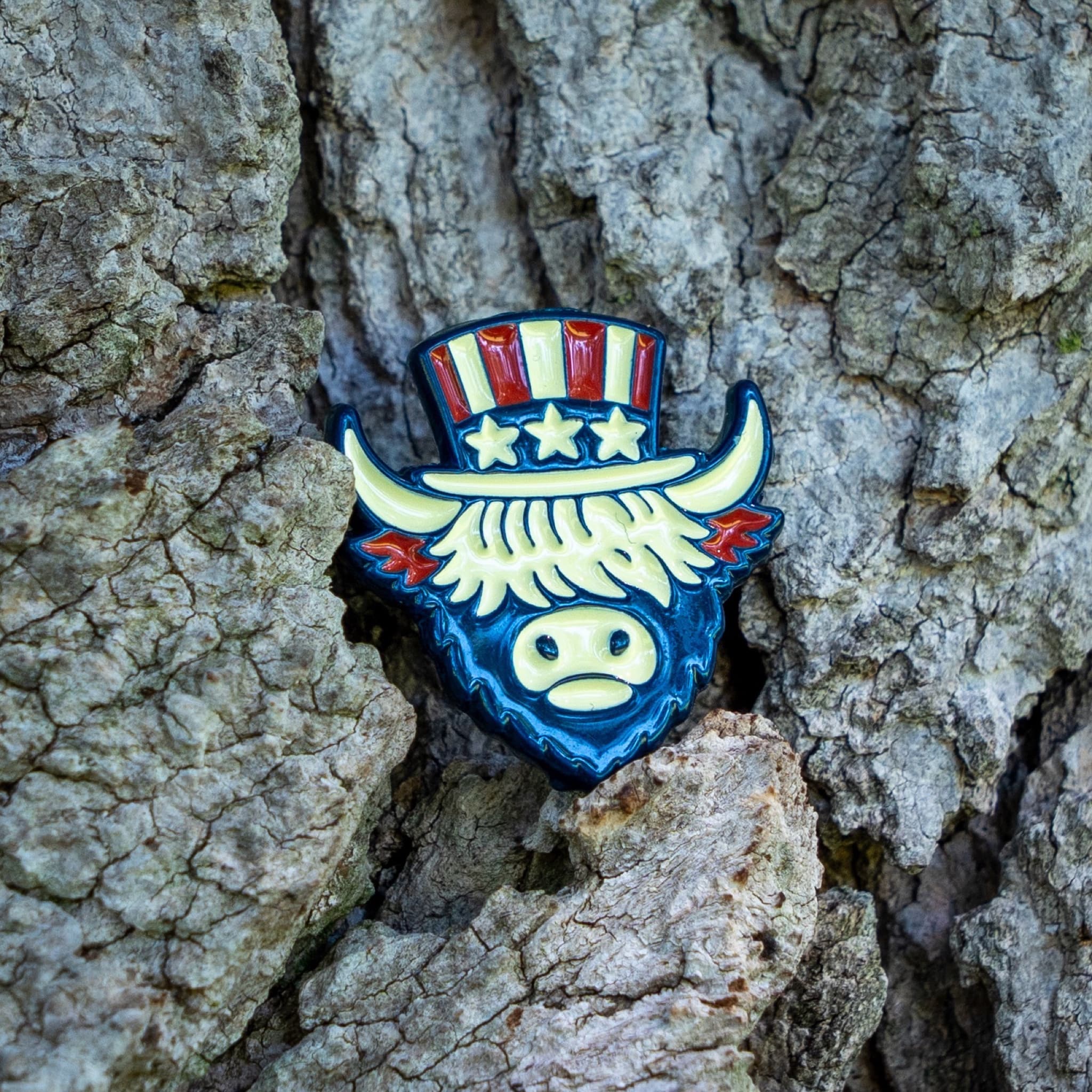 A patriotic buffalo head enamel pin with a top hat design, featuring red, white, and blue colors, photographed against a textured tree bark background.