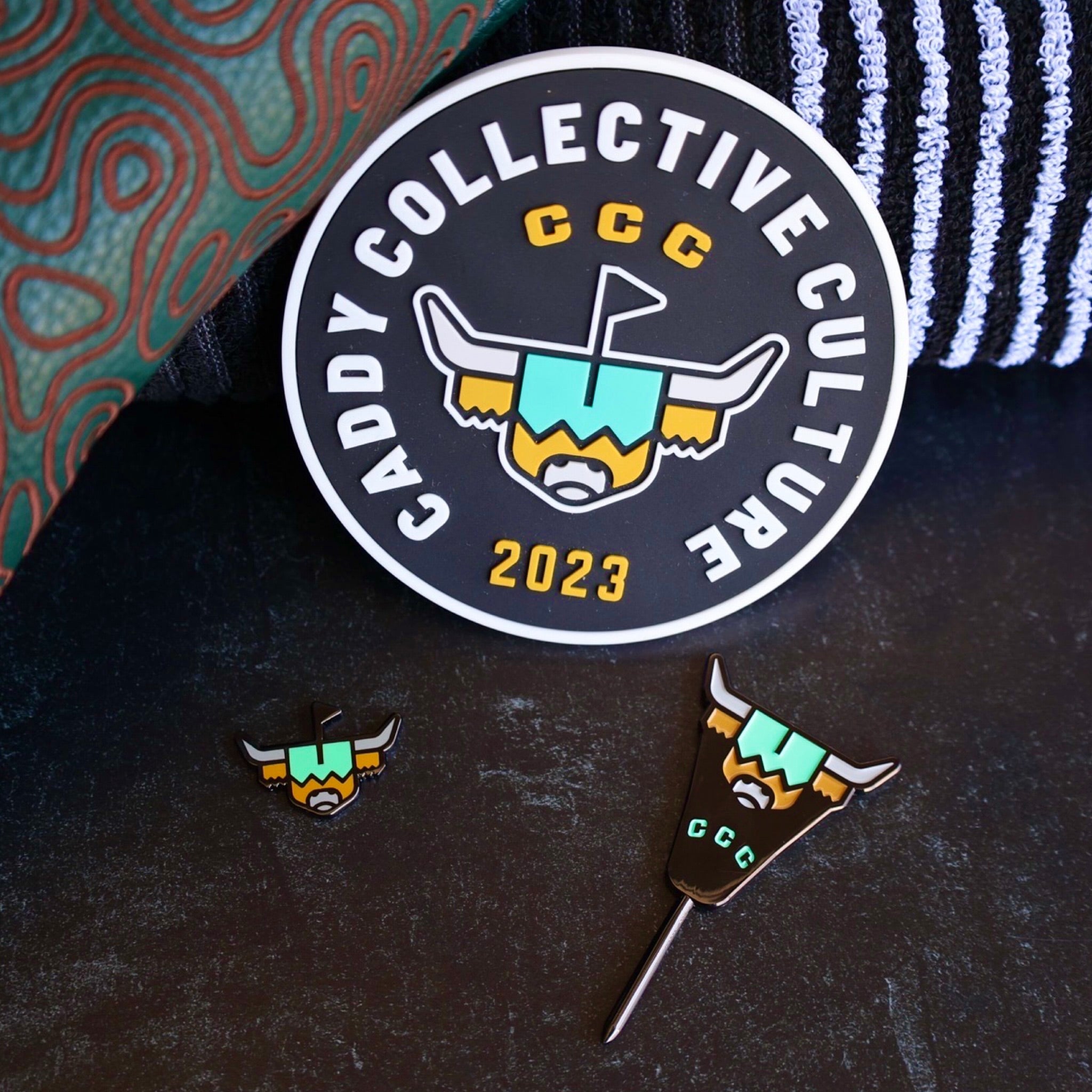 Flat lay detail of Looper’s Field Kit golf accessories including CCC practice disc, ball marker and divot tool on black striped towel.