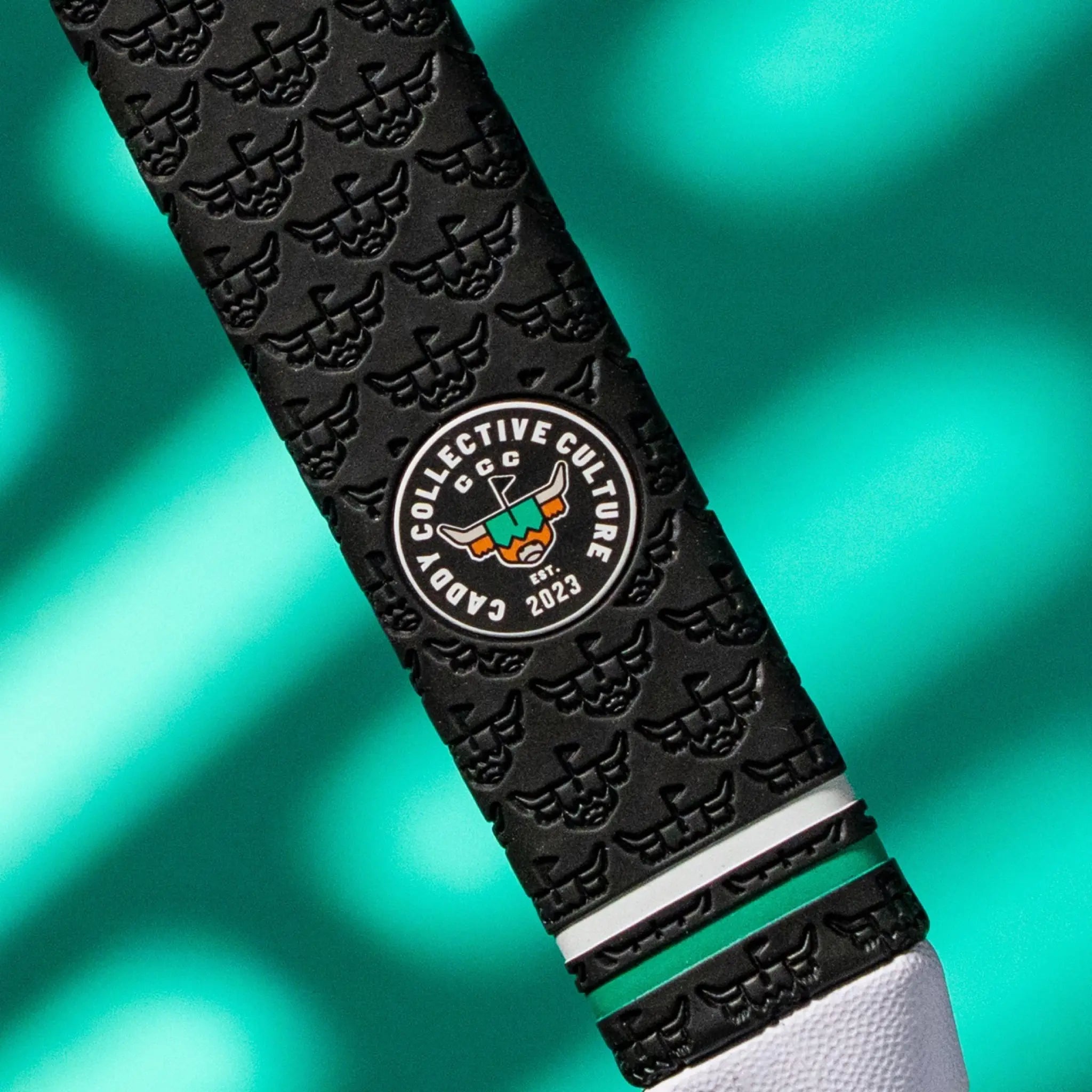 Close-up of a black Garsen putter grip showing the Caddy Collective Culture badge, repeating embossed caddy pattern, green accent ring, and white end cap.