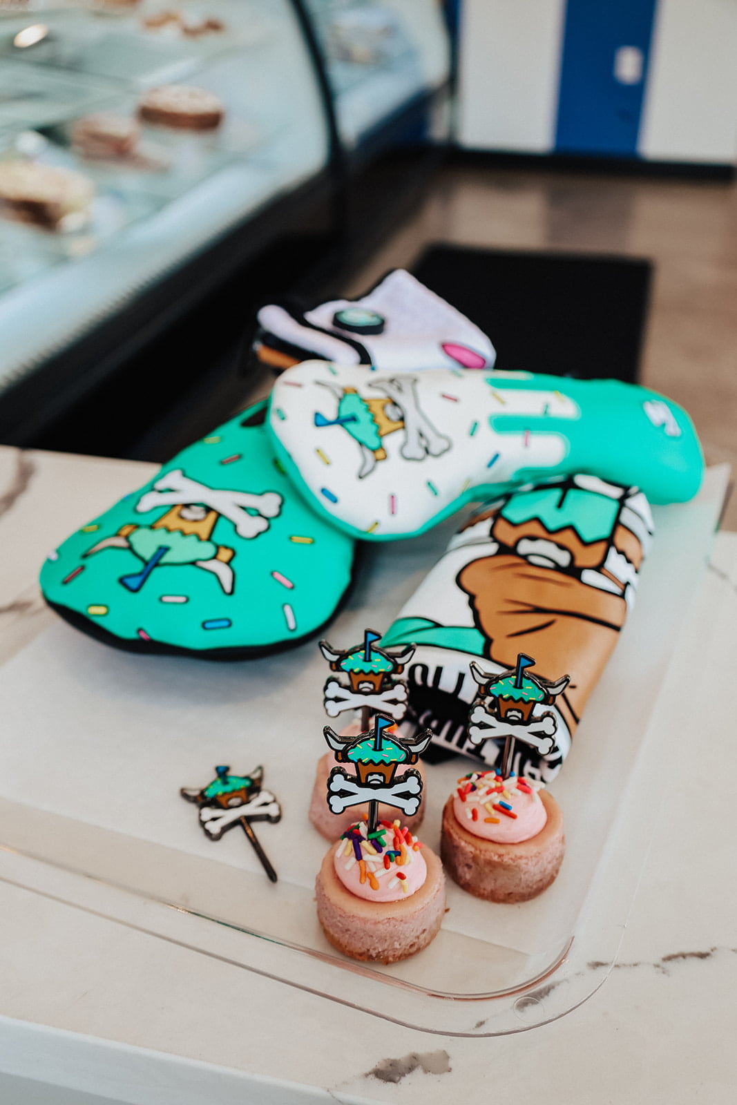 Johnny Cupcakes x Caddy Collective Culture golf collection with headcovers, towel, and divot tools displayed with pink cupcakes topped with sprinkles.