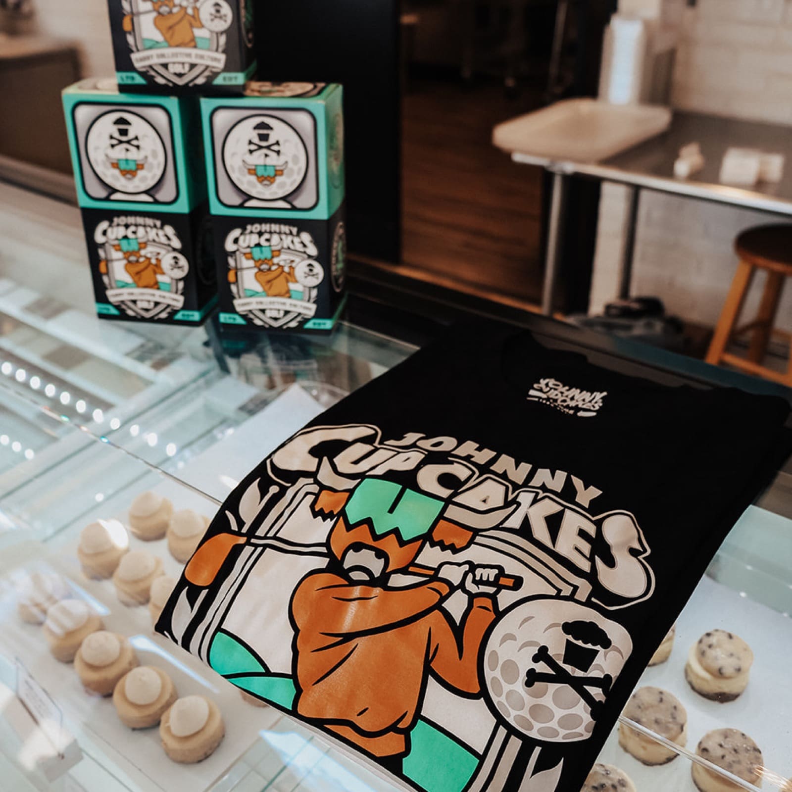 Johnny Cupcakes x Caddy Collective Culture black golf shirt with cupcake golfer design, displayed with packaging boxes and bakery treats.