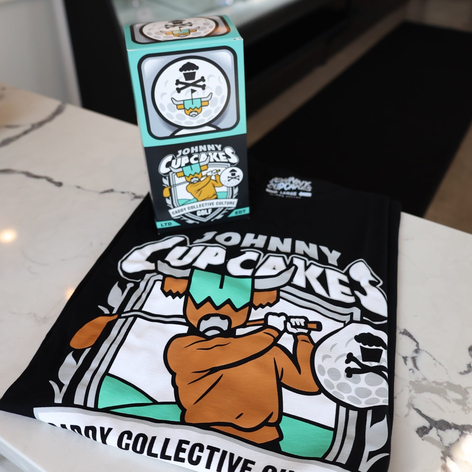 Johnny Cupcakes x Caddy Collective Culture black golf shirt with cupcake golfer design, displayed with limited edition packaging box.