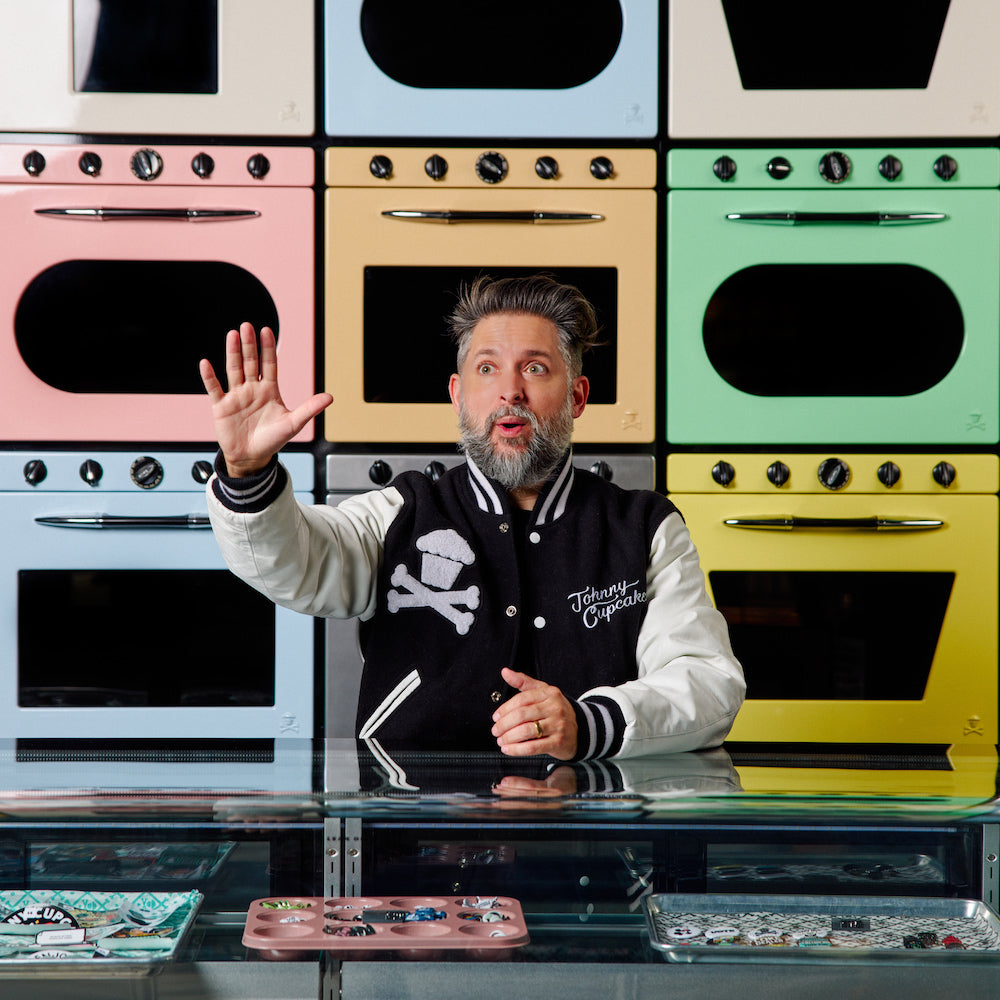 Johnny Cupcakes standing in front of pastel oven wall wearing branded varsity jacket — image used for CCC x Johnny Cupcakes limited-edition collaboration story.