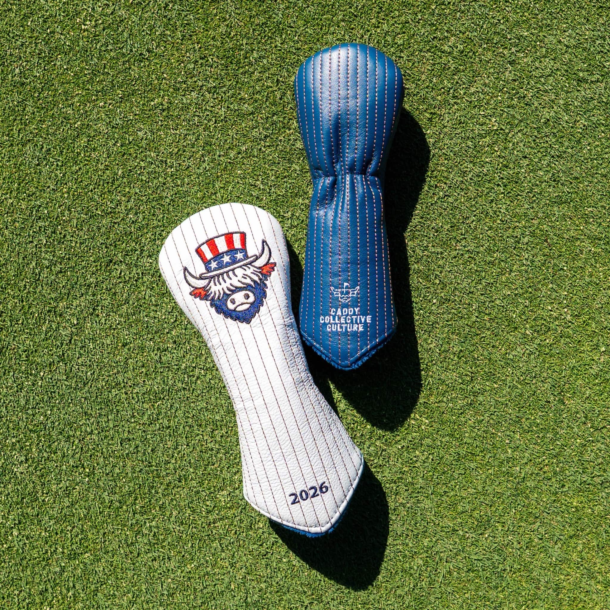 Two golf club headcovers placed on green grass: a white pinstripe cover with a buffalo mascot logo and red, white, and blue patriotic hat graphic, and a blue cover with white text reading 'Cade Collective Golf.'.