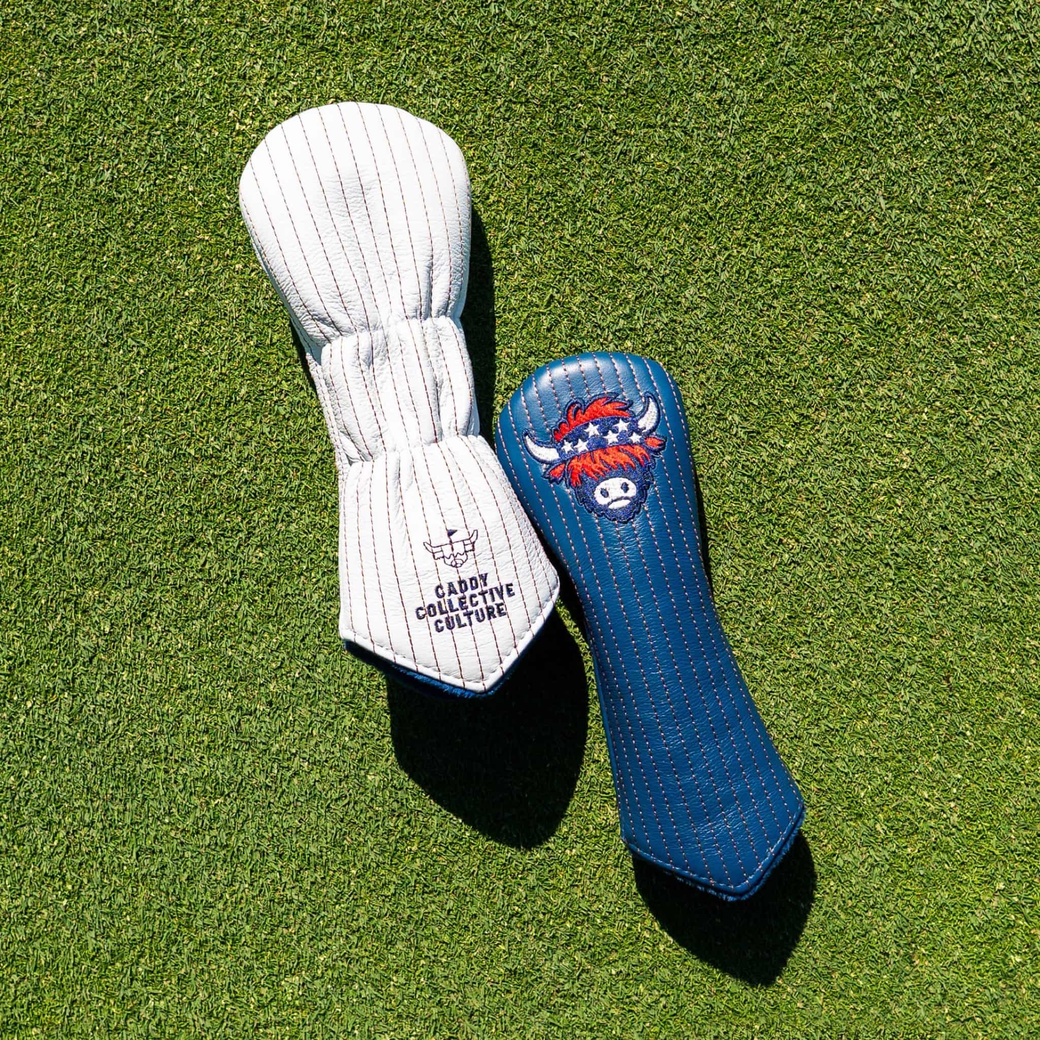 Two golf club headcovers placed on green grass: one white with thin dark pinstripes and the text 'Caddy Collective Culture,' and one blue with a red, white, and blue highlands cattle logo graphic.