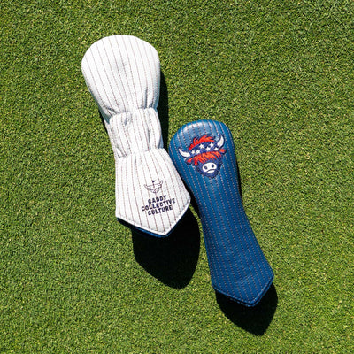 Two golf club headcovers placed on green grass: one white with thin dark pinstripes and the text 'Caddy Collective Culture,' and one blue with a red, white, and blue highlands cattle logo graphic.