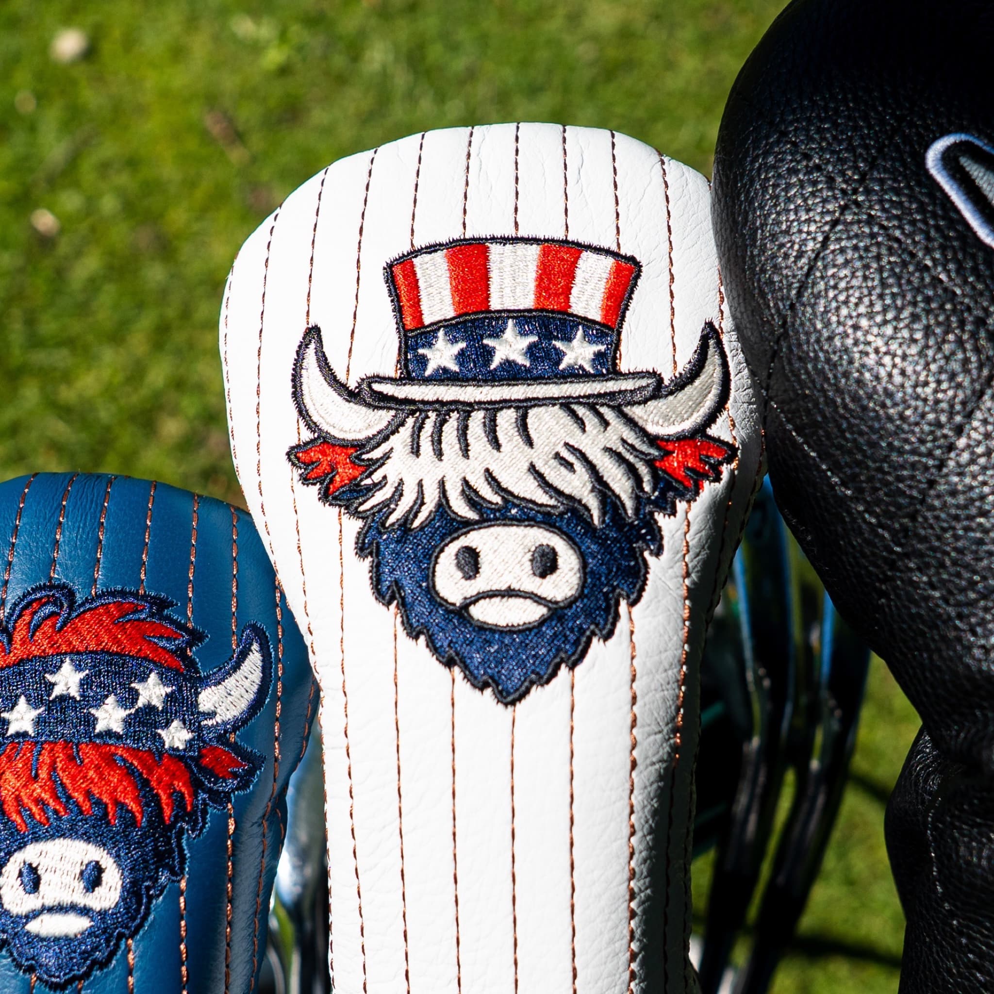 Close-up of a white pinstriped golf club headcover with an embroidered buffalo mascot wearing a red, white, and blue top hat, with part of a blue matching headcover and a black textured club head visible beside it on grass.