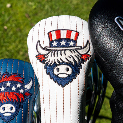Close-up of a white pinstriped golf club headcover with an embroidered buffalo mascot wearing a red, white, and blue top hat, with part of a blue matching headcover and a black textured club head visible beside it on grass.