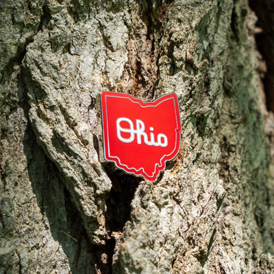 A red enamel pin shaped like the state of Ohio with the word 'Ohio' in white script, attached to the side of a textured tree trunk.