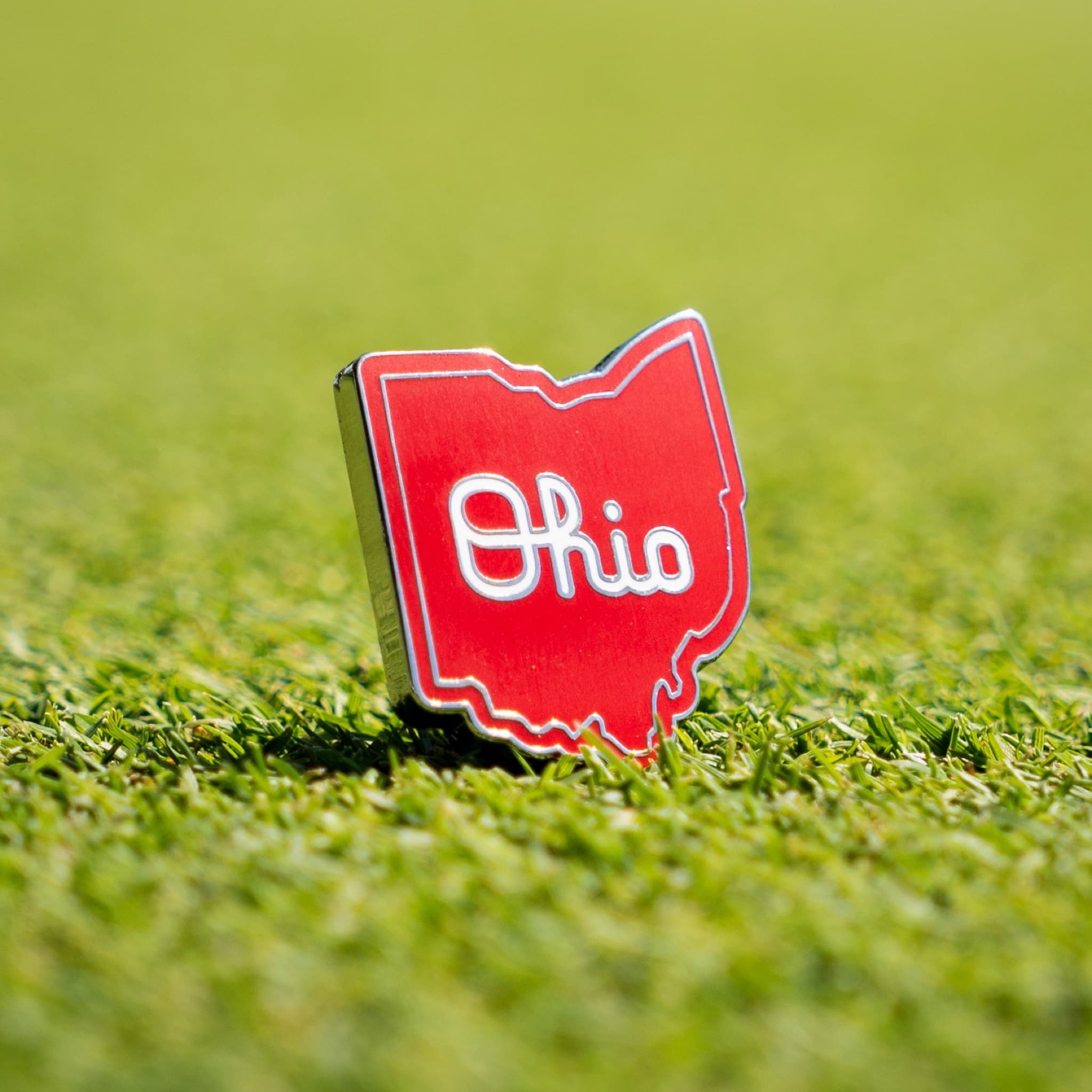 A red enamel pin shaped like the state of Ohio with the word 'Ohio' in white script, standing upright on a green grass surface.