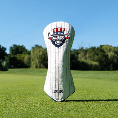 A white pinstripe golf club headcover standing upright on a green grass field under a clear blue sky. The cover features an embroidered highland cow mascot wearing a red, white, and blue top hat with stars, and the year '2026' printed near the bottom.