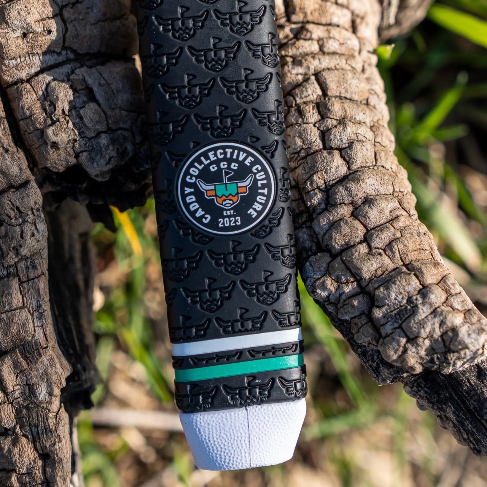 Vertical close-up of a black Garsen putter grip with repeating embossed logos, Caddy Collective Culture badge, green accent ring, and white end cap against rustic wood.