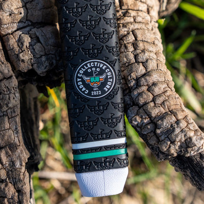 Vertical close-up of a black Garsen putter grip with repeating embossed logos, Caddy Collective Culture badge, green accent ring, and white end cap against rustic wood.