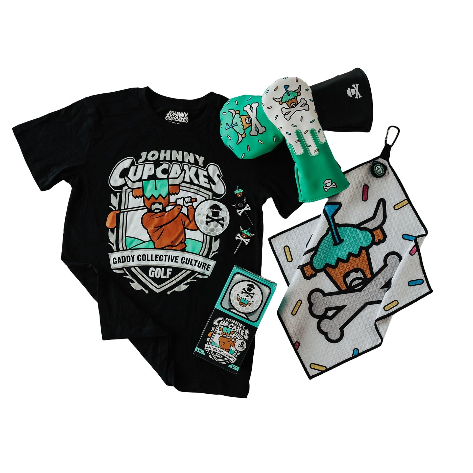 Johnny Cupcakes x Caddy Collective Culture golf collaboration flatlay featuring t-shirt, driver headcovers, towel, divot tool, and collectible box.