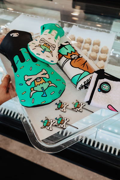 Johnny Cupcakes x Caddy Collective Culture full golf collection including driver and fairway headcovers, divot tools, towel, and shirt displayed on bakery tray.