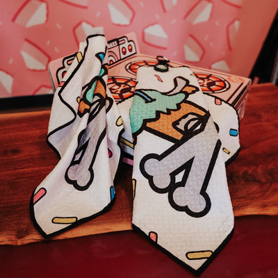 Johnny Cupcakes x Caddy Collective Culture golf towel with cupcake and crossbones design, displayed on wood surface with bakery oven box and cheesecake wall backdrop.