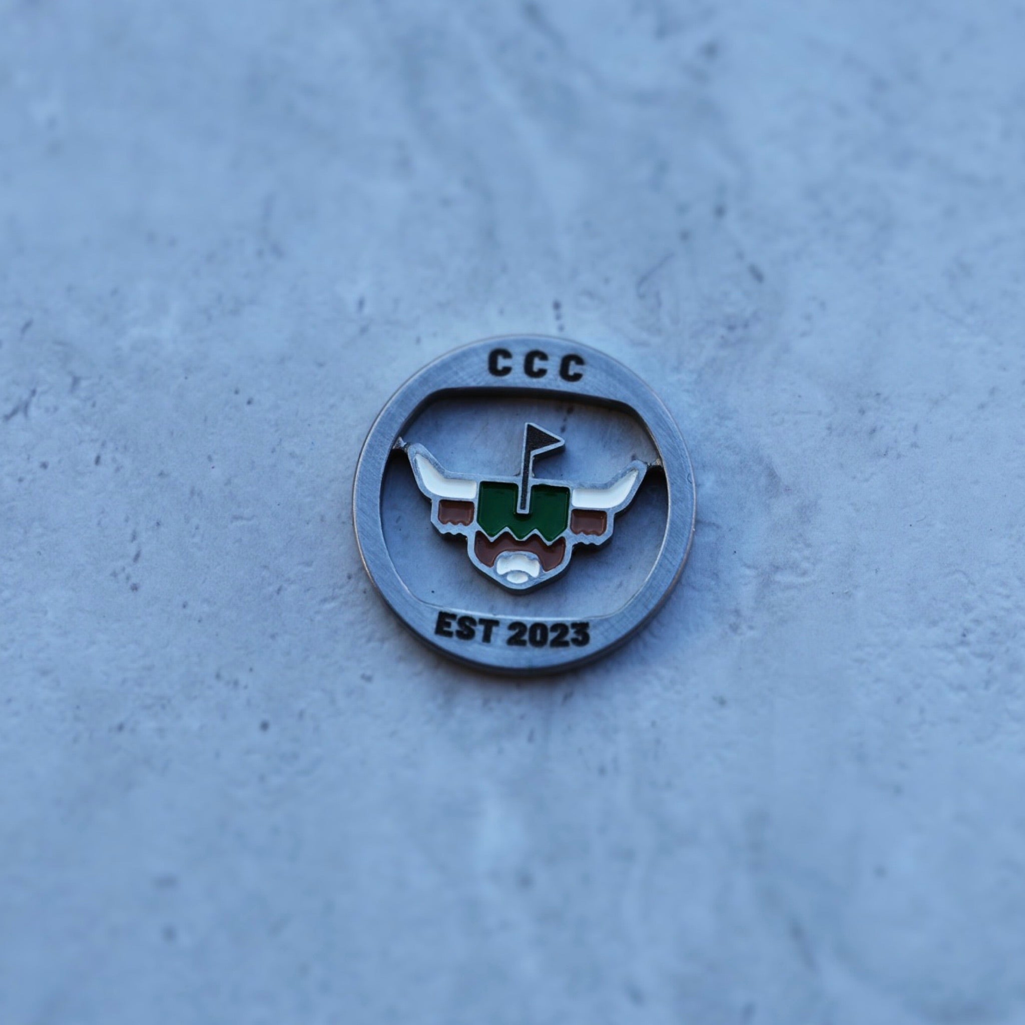 First Cut CCC Mark ball marker photographed on textured stone surface, showing CNC-milled metal and enamel logo detail.