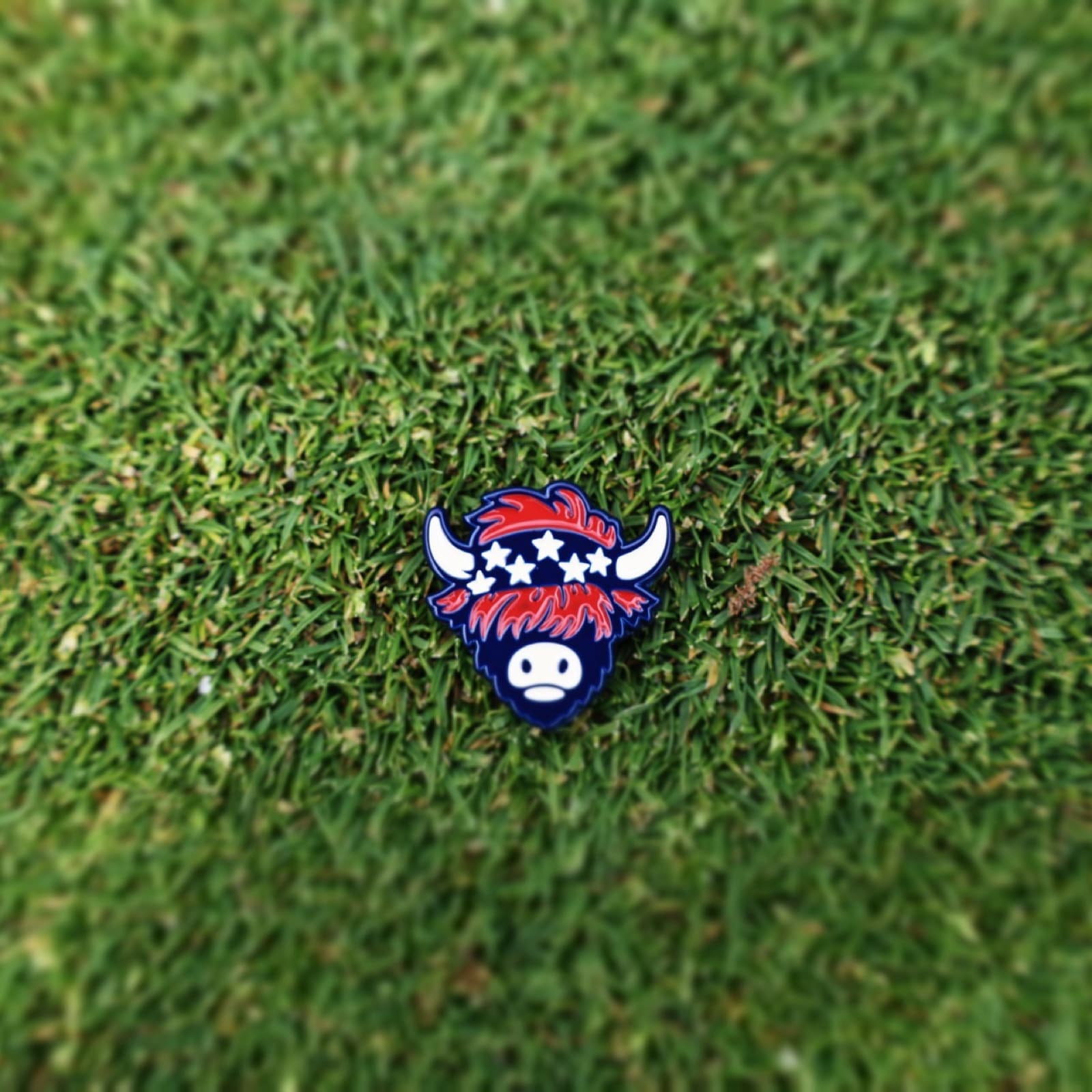 Patriotic golf ball marker by Caddy Collective Culture resting on putting green grass.