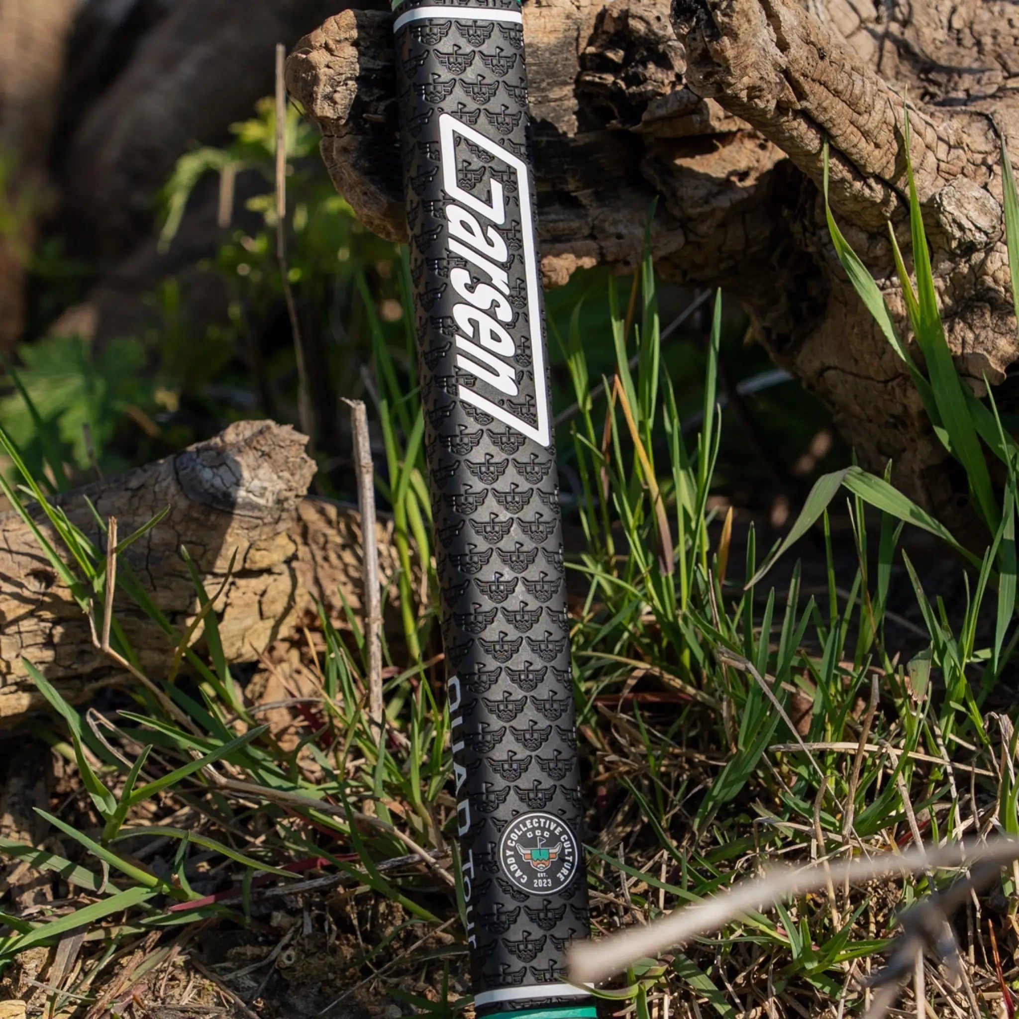 Outdoor lifestyle photo of a black Garsen putter grip with white Garsen branding, Caddy Collective Culture badge, and embossed pattern near grass and weathered wood.