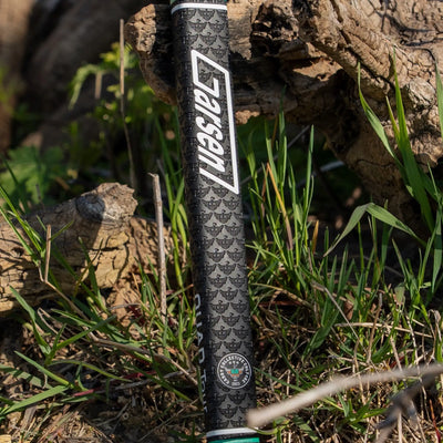 Outdoor lifestyle photo of a black Garsen putter grip with white Garsen branding, Caddy Collective Culture badge, and embossed pattern near grass and weathered wood.