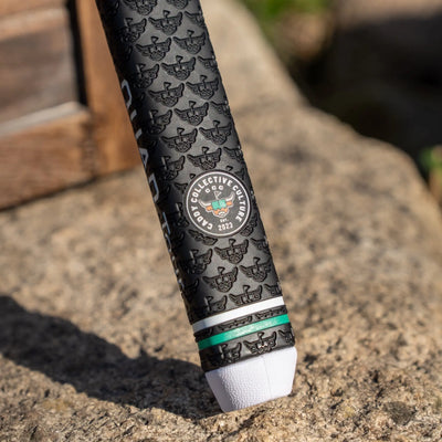 Close-up of a black Garsen putter grip with a Caddy Collective Culture logo badge, embossed pattern, green accent ring, and white end cap resting on a rock.