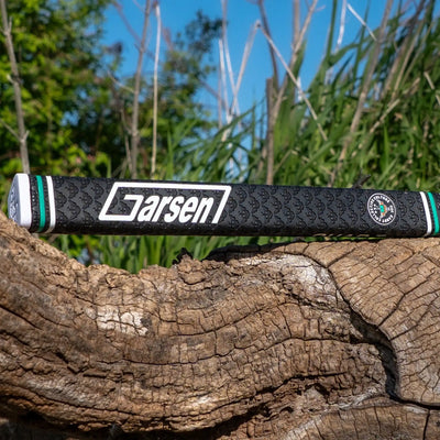 Full product shot of a black Garsen putter grip resting on weathered wood, featuring a white Garsen logo, Caddy Collective Culture badge, and green accent rings.