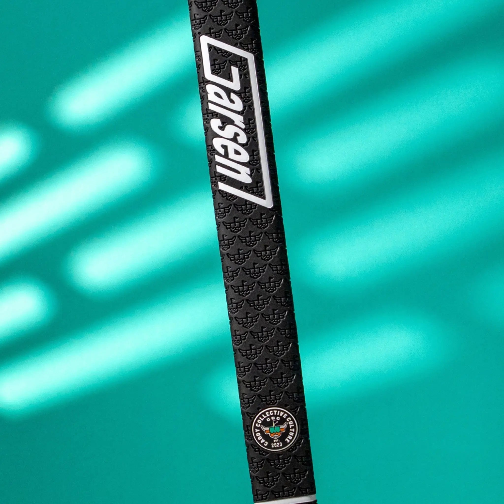 Angled view of a black Garsen QUAD Tour putter grip featuring the white Garsen logo, Caddy Collective Culture badge, and embossed texture on a teal background.